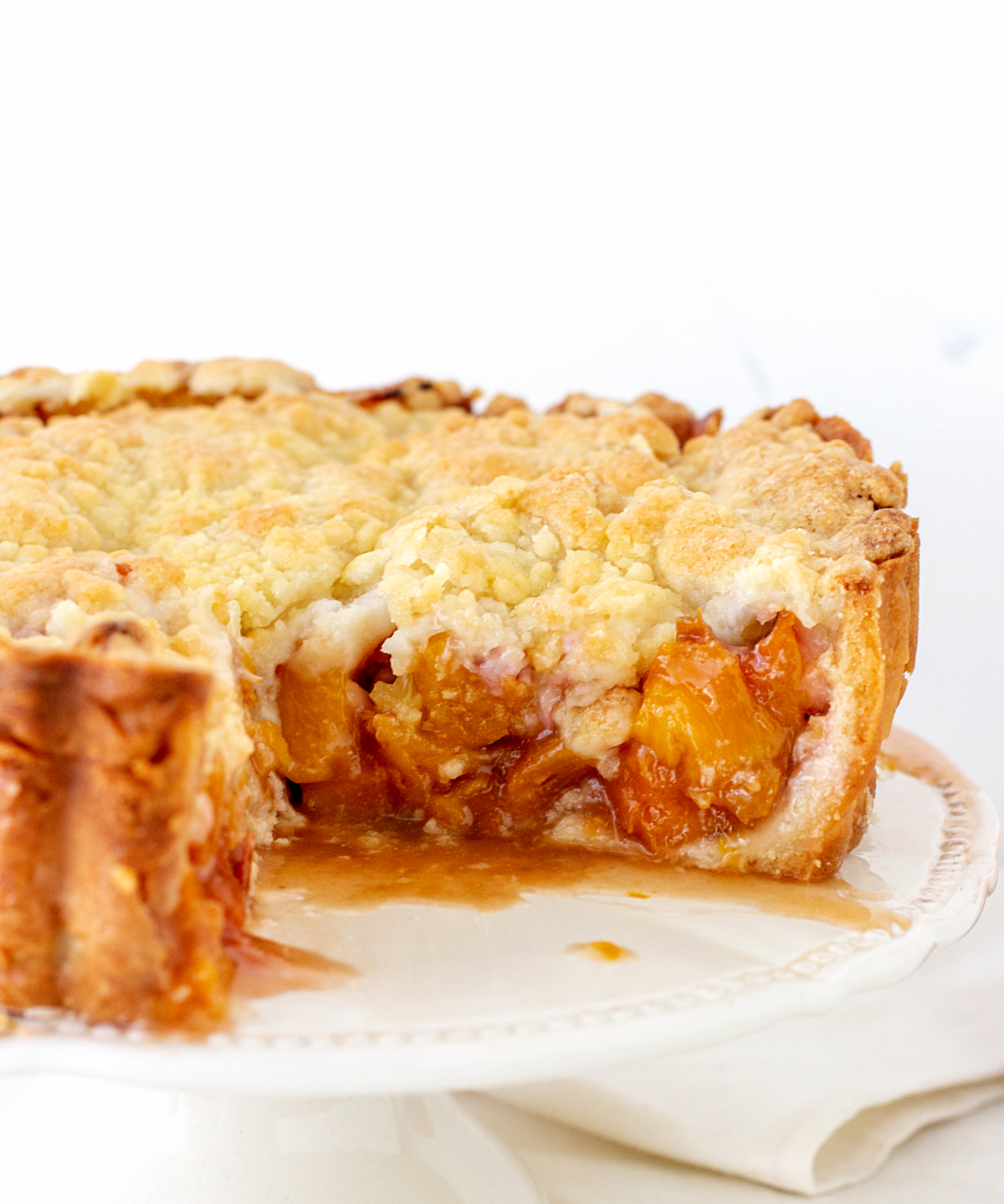 Peach crumble pie on a white cake stand, piece missing. White background.