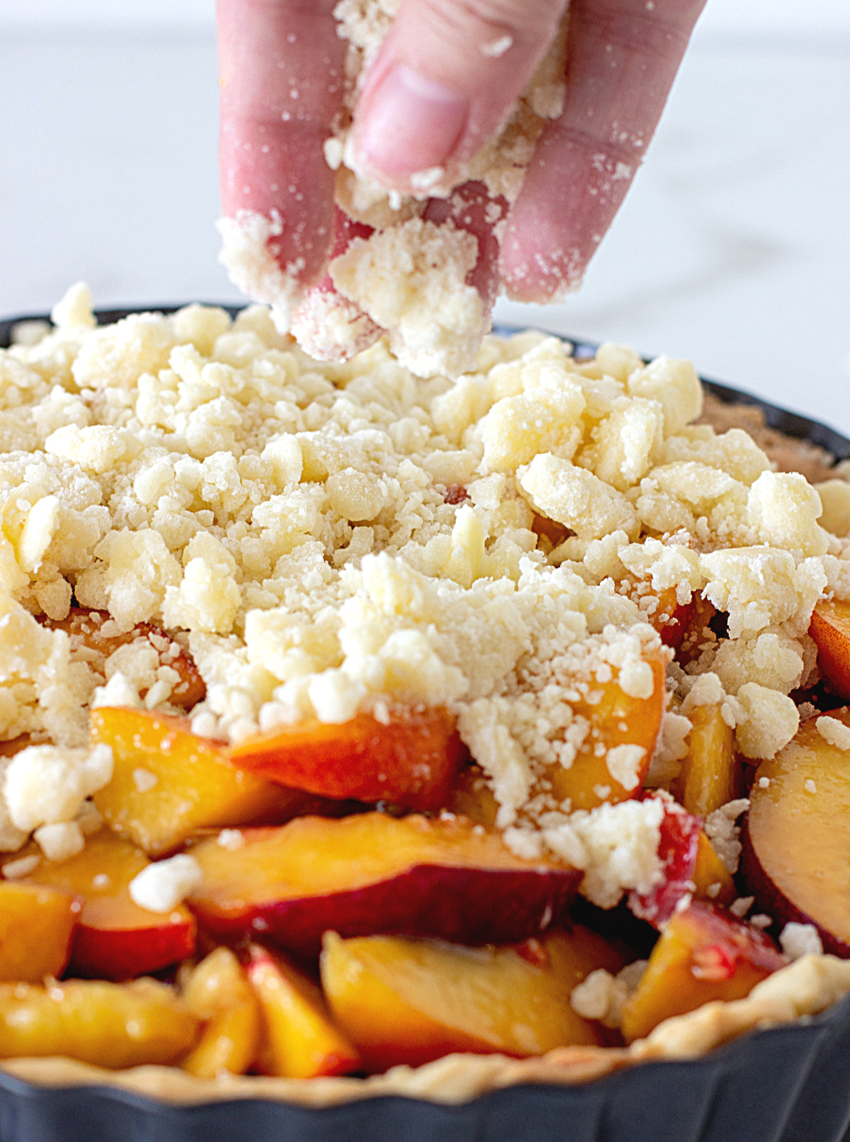 Hand adding crumble to peaches in pie metal pan.