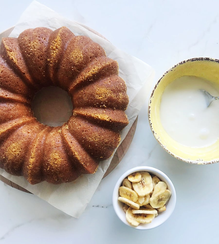 On a white surface, a plain bundt cake, bowl with glaze, and bowl with banana chips.