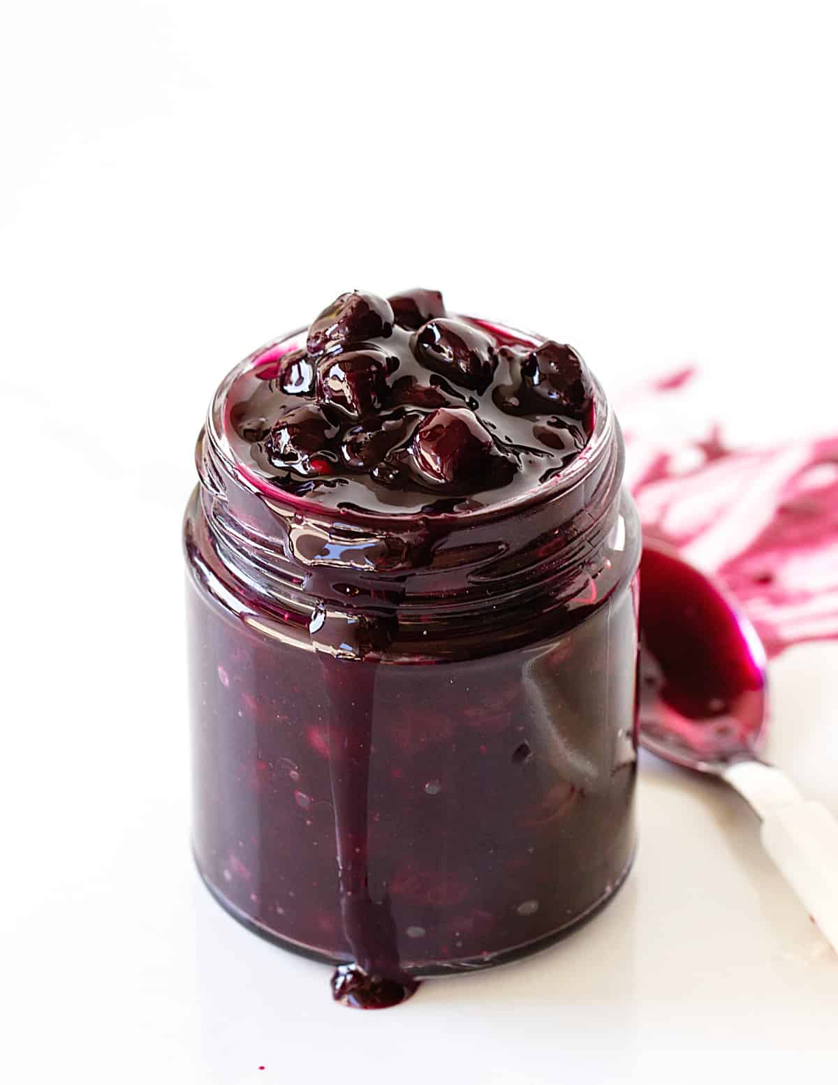 Glass jar with blueberry sauce, drip and spoon, white background