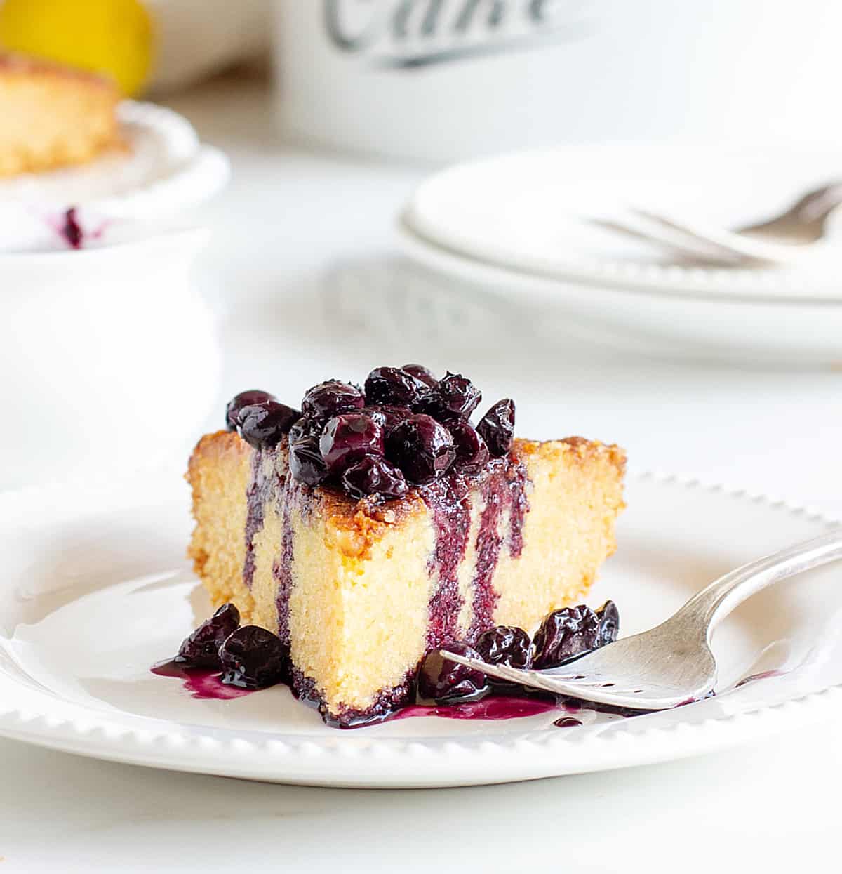 Slice of lemon cake with blueberry sauce on white plate, silver fork, white props in the background.