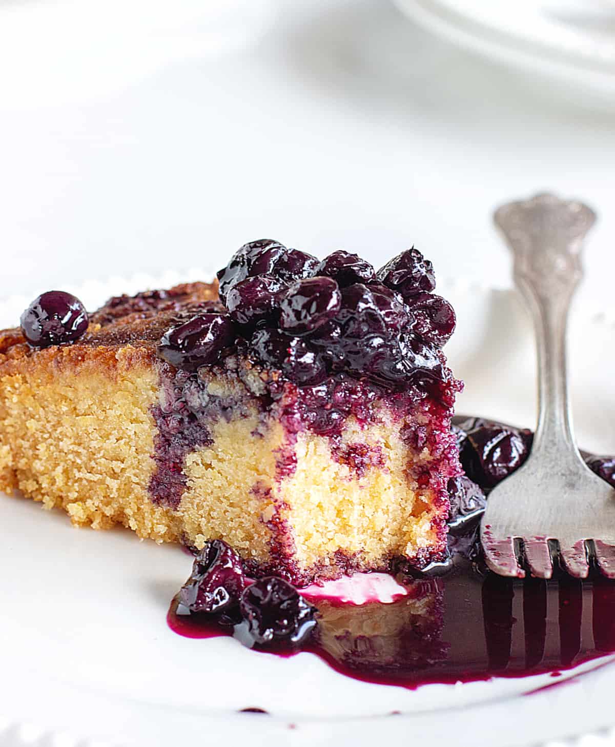 Eaten slice of polenta cake with pool of blueberry sauce, white plate, silver fork.