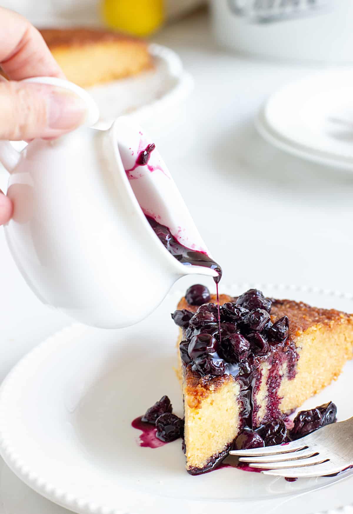 Pouring blueberry sauce on slice of yellow cake from white saucer, white plate