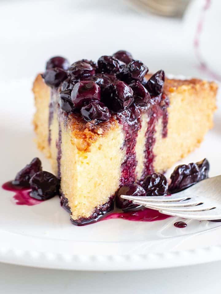 Slice of lemon cake with blueberry sauce on white plate, silver fork, white background.