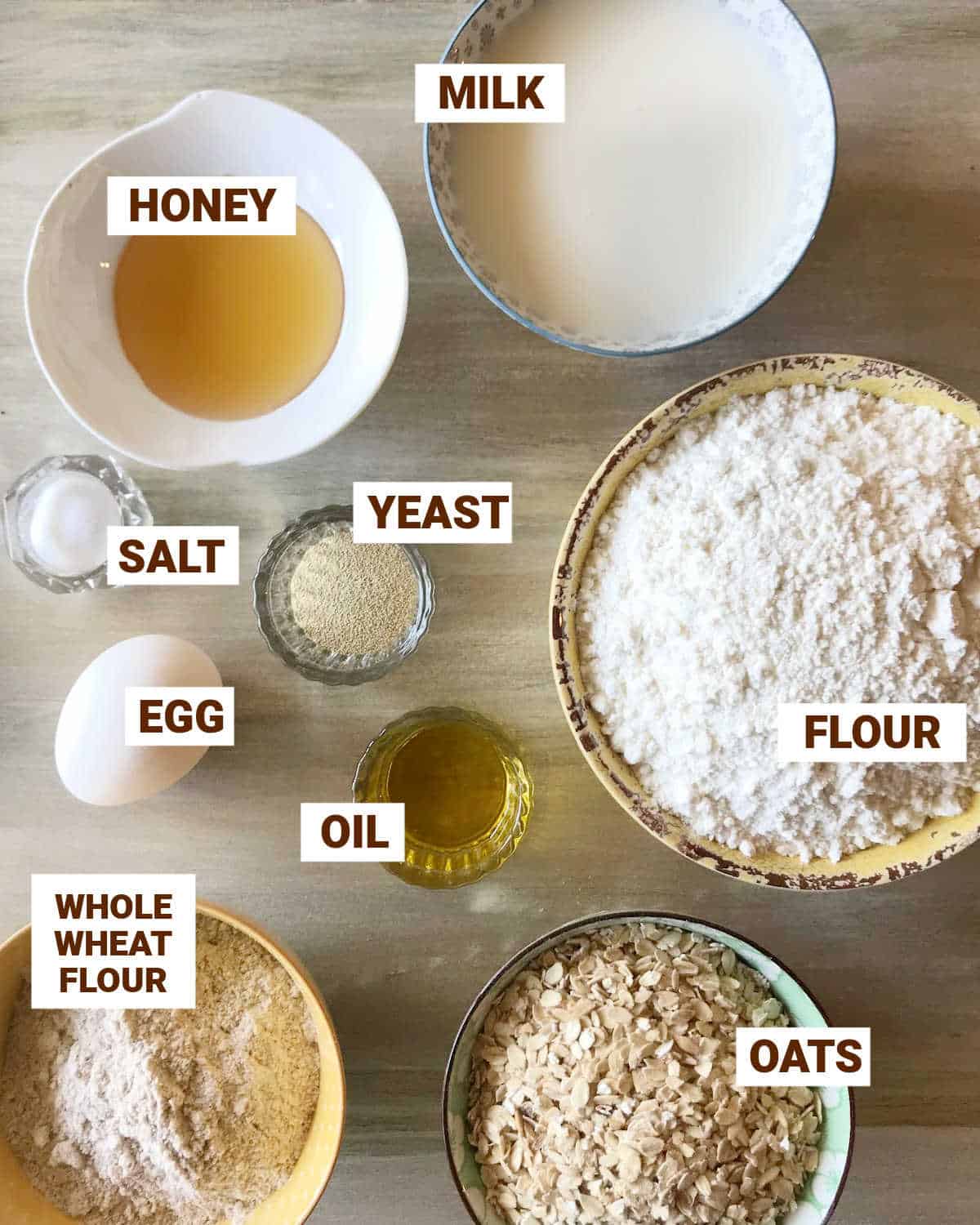 Whole wheat oatmeal bread ingredients in bowls on a beige surface including milk, honey, egg, yeast, oil, flours, salt.