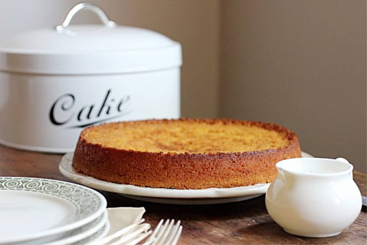 Whole short Lemon Cake on wooden table, stack of plates, saucer, cake tin in the background.