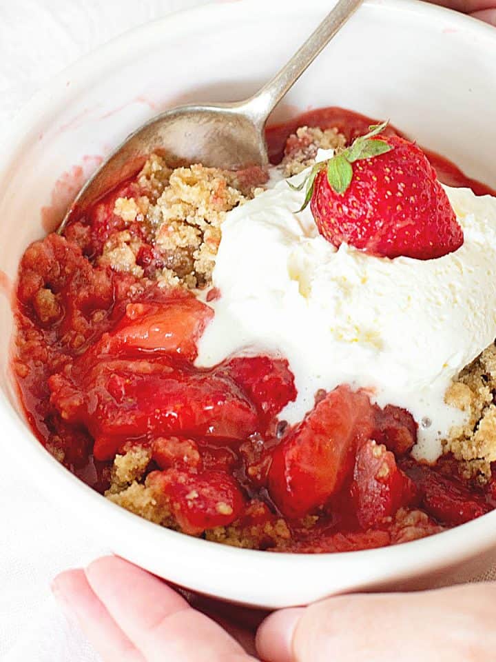 Hands holding white bowl with strawberry crisp and ice cream, silver spoon