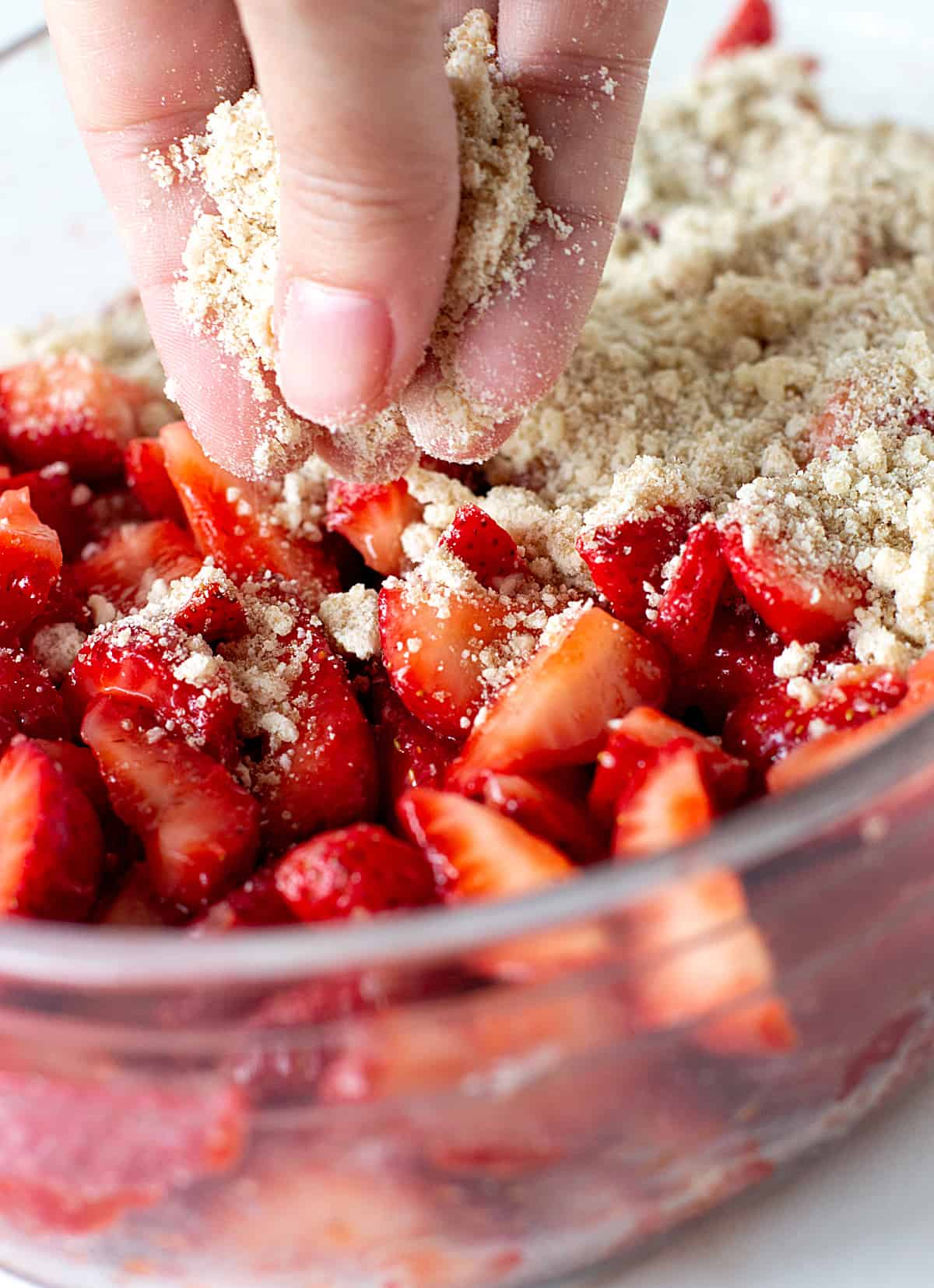 Hand adding crumble topping to glass dish with strawberries