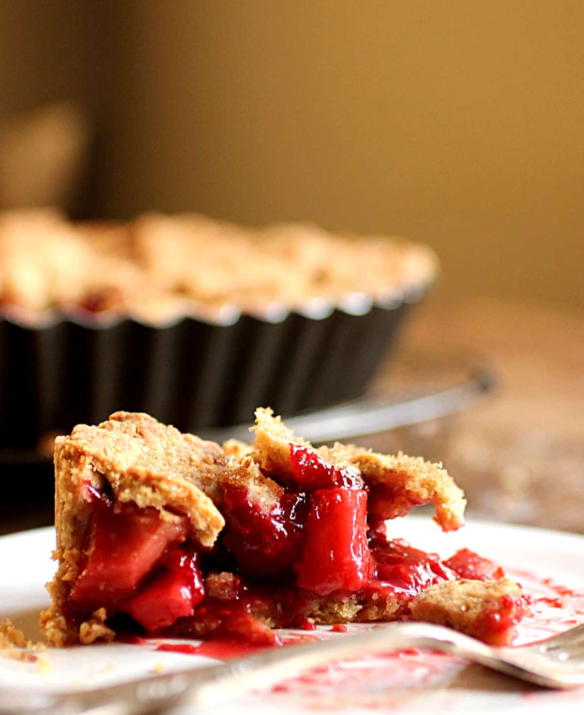 Slice of apple raspberry pie on white plate, pie pan and wooden table on the background.