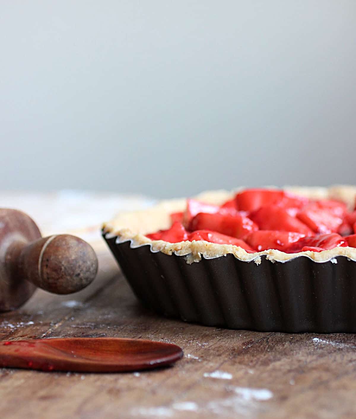 Metal pie pan with red filling on wooden table, rolling pin and wooden spoon.