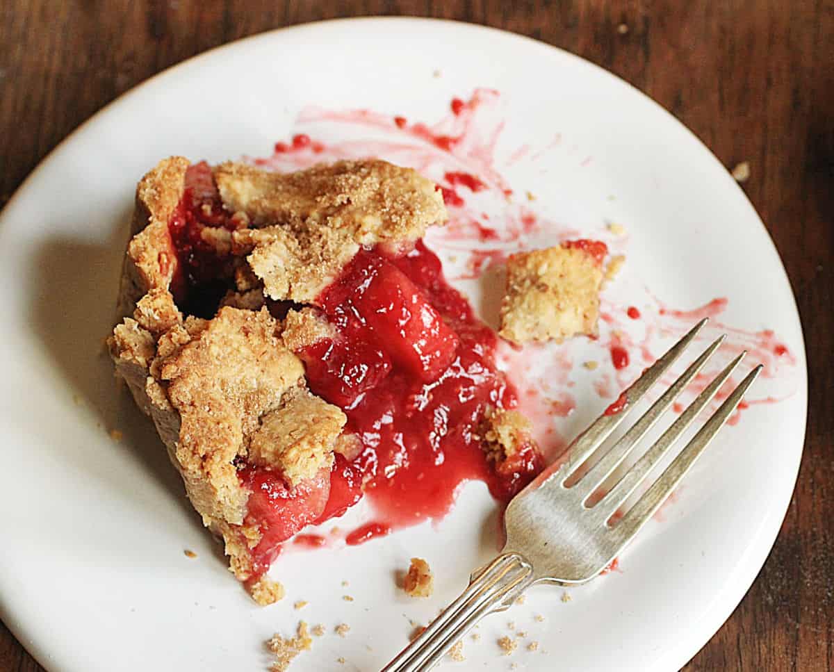 Slice of raspberry apple pie on white plate, silver fork, wooden table.