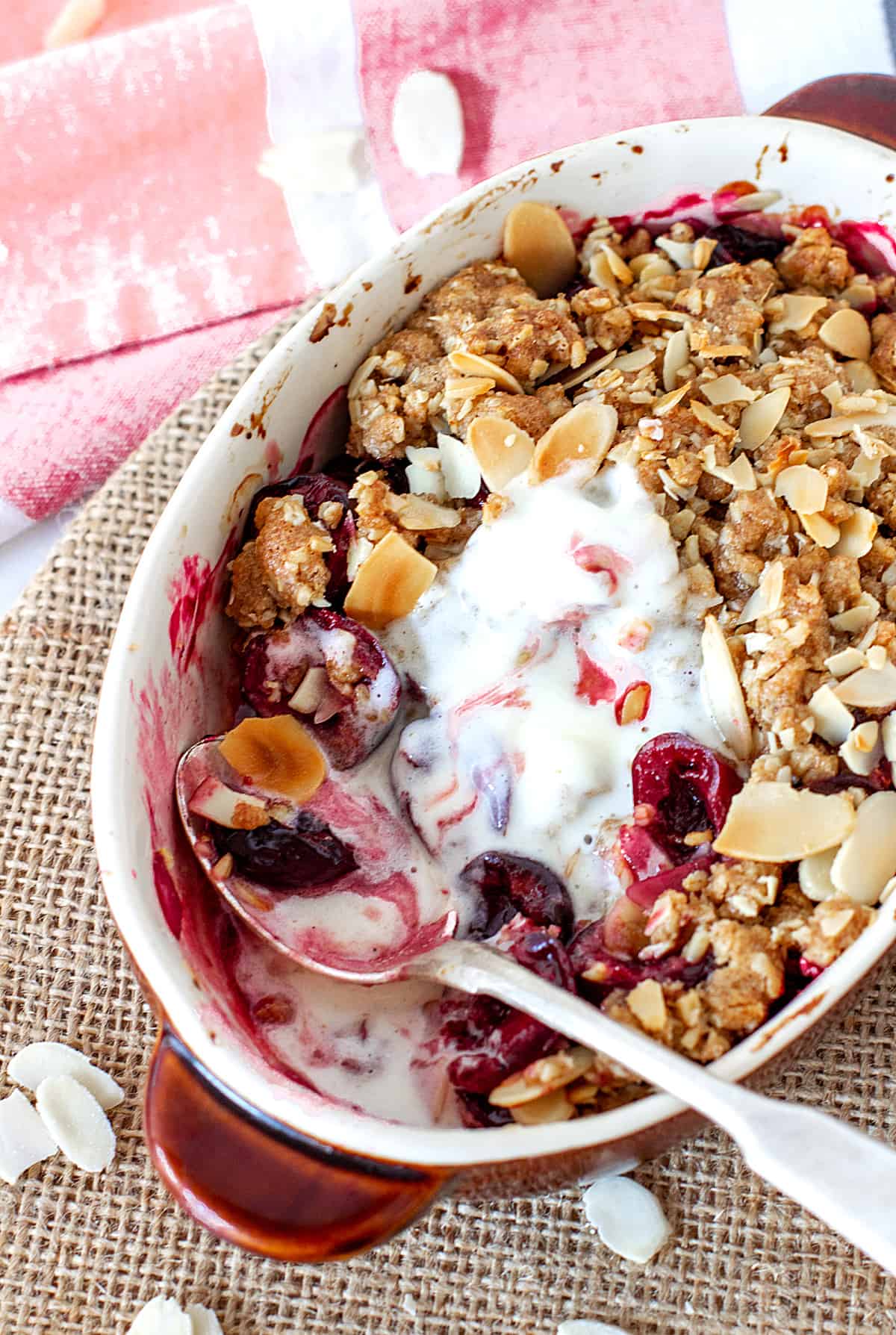 Individual oval dish with cherry crisp, silver spoon, pink kitchen towel and burlap surface