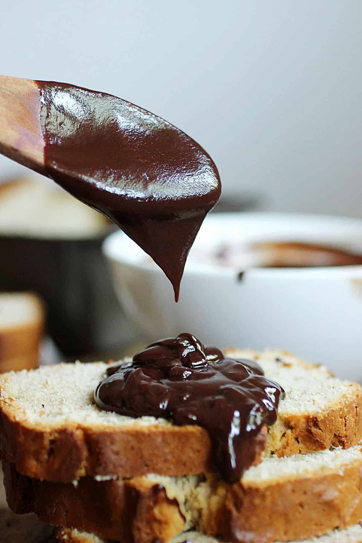 Wooden spoon drizzling chocolate sauce on stack of quick bread slices, white bowl in the background.