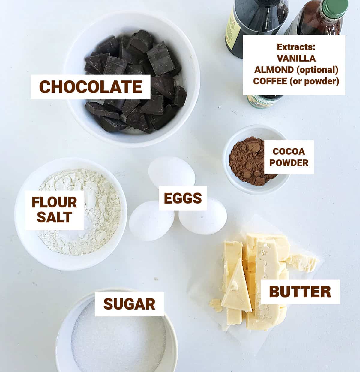 White surface with bowls containing brownie ingredients, like eggs, extract bottles, image with text.