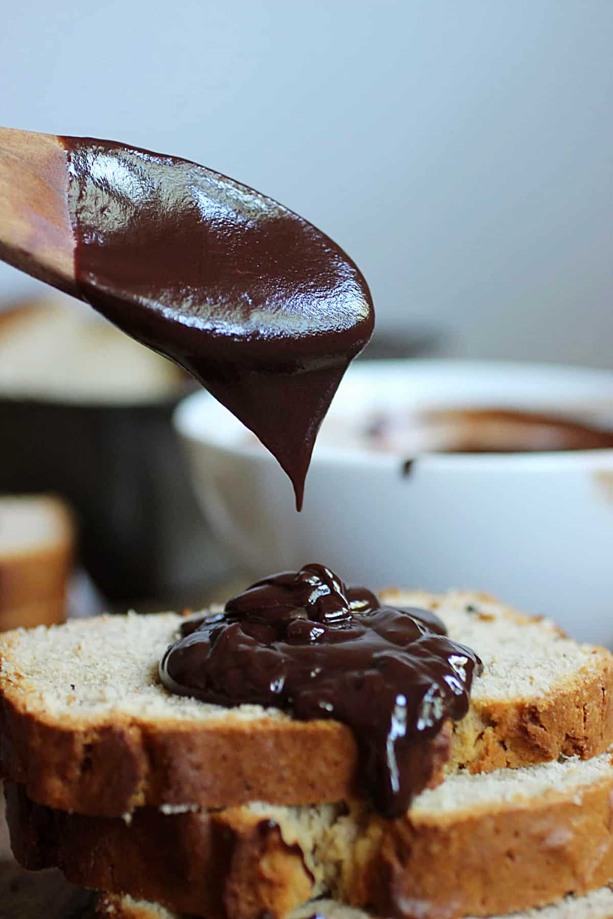 Wooden spoon drizzling chocolate sauce on stack of quick bread slices, white bowl