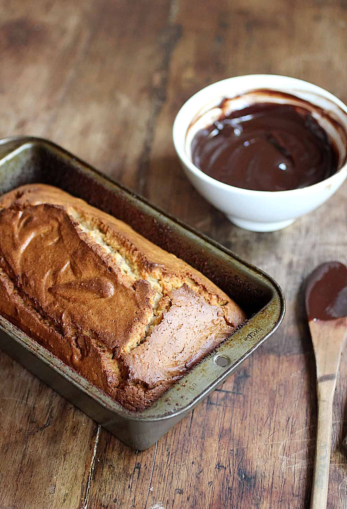 Loaf of quick bread in metal pan, bowl with chocolate sauce, wooden table.