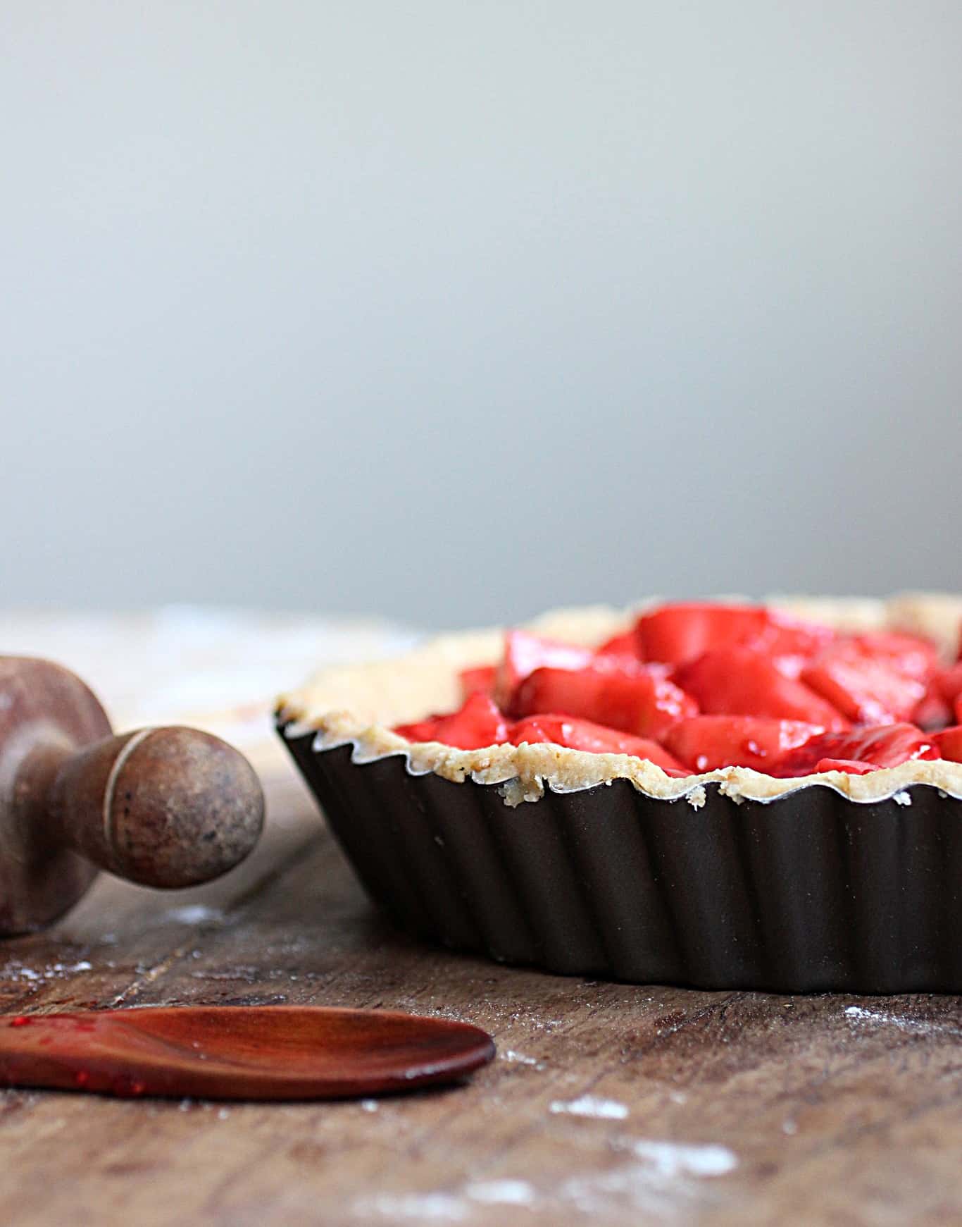 Metal pie pan with red filling on wooden table, rolling pin and wooden spoon