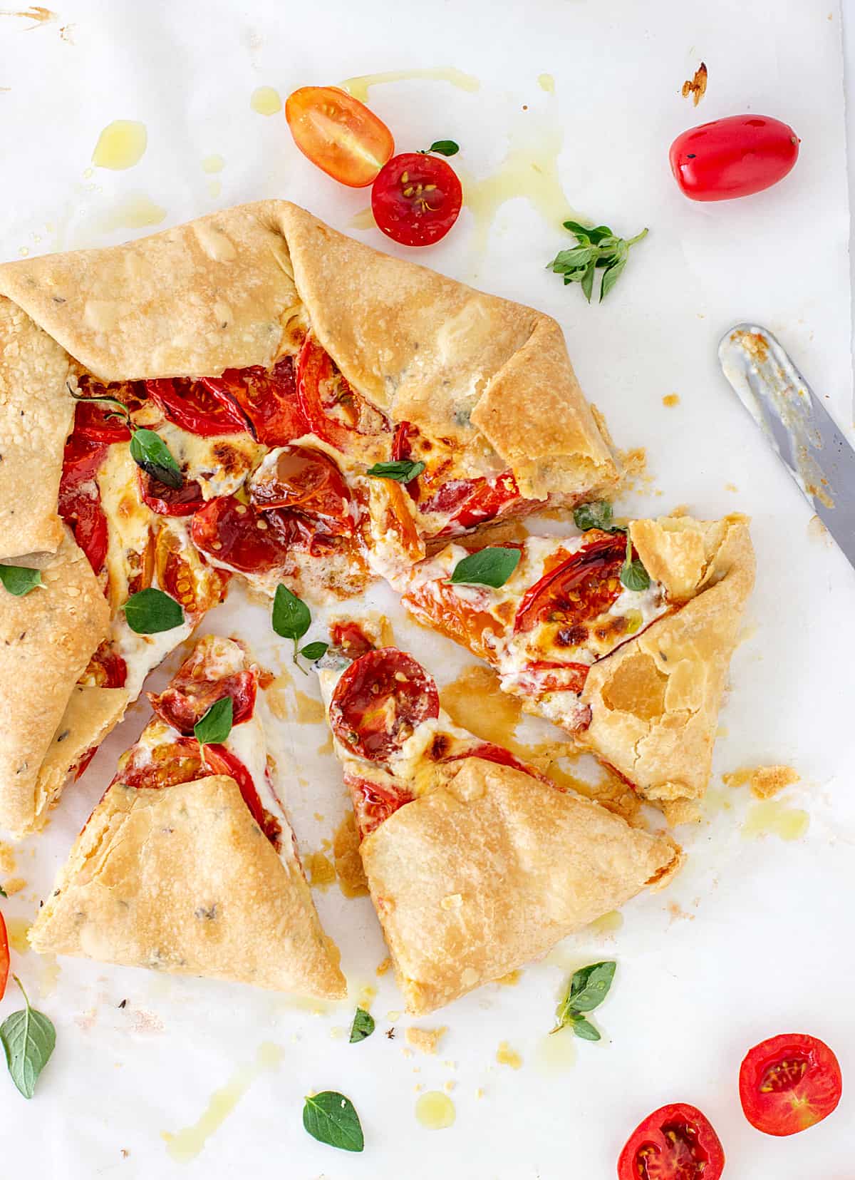 Slices and half piece of tomato galette on white surface, loose herbs