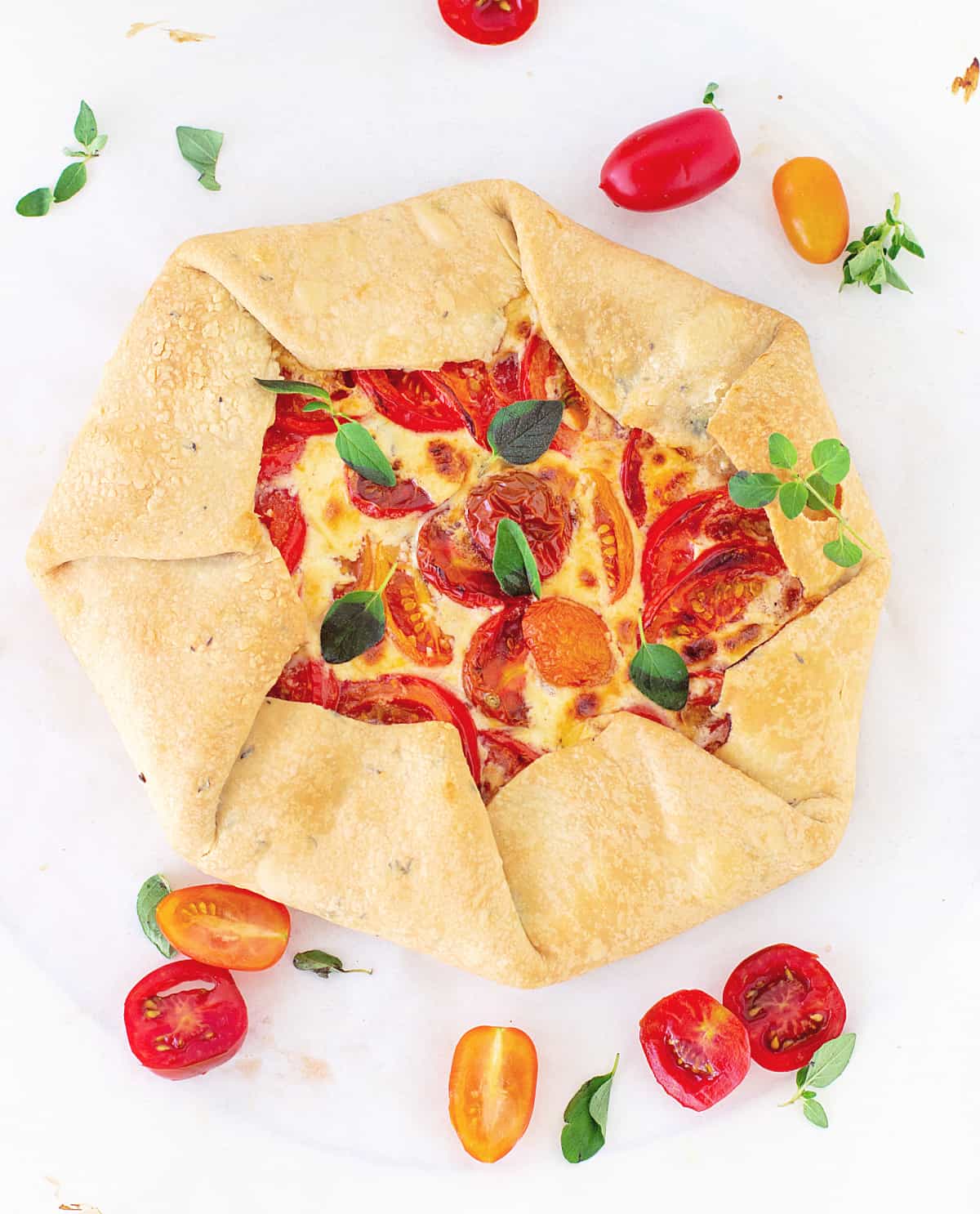 Top view of whole tomato galette on white surface, loose cherry tomatoes and herbs around it.