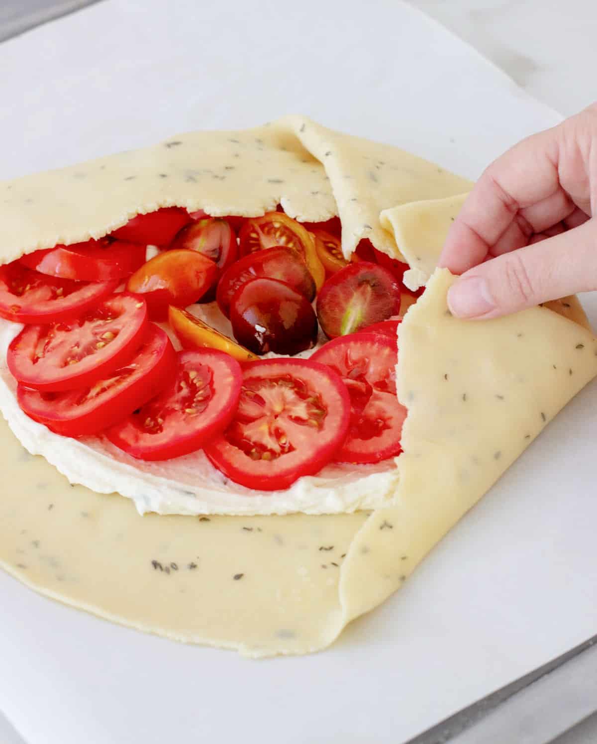 Hand folding pie crust over tomato filling in galette placed on white parchment paper.