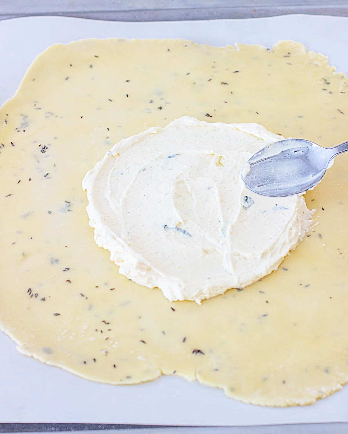 Spreading cream cheese mixture with spoon on round of pie crust on white parchment paper.