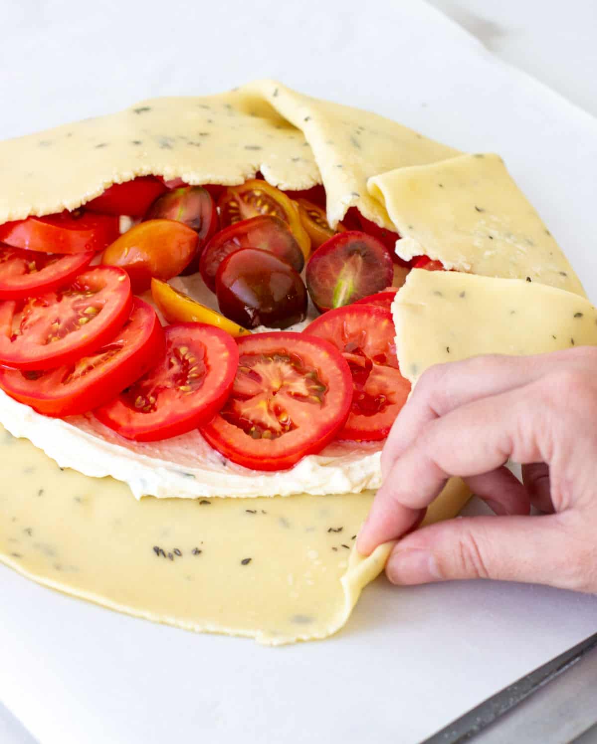 Lifting edge of herbed pie crust with hand to encase tomato filling. Dough on white parchment paper.