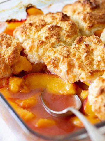 Glass dish with half eaten peaches in syrup, biscuit topping and silver spoon.