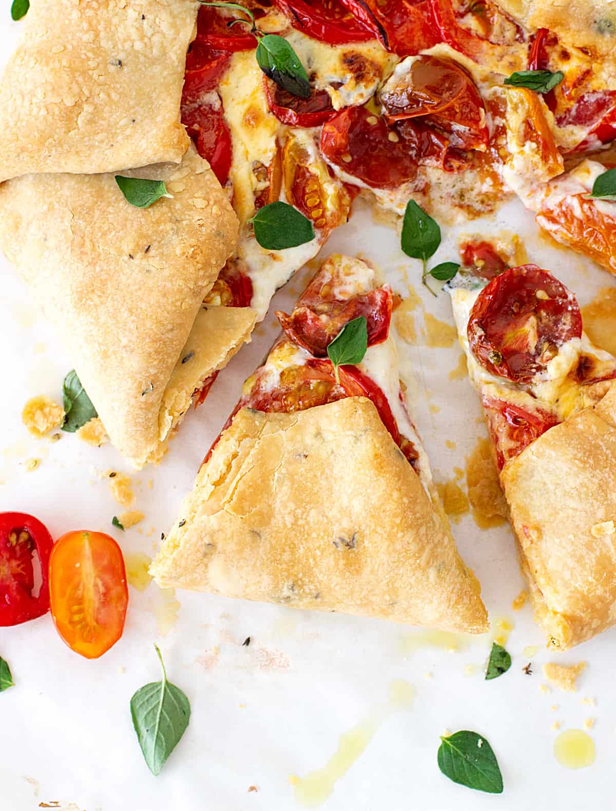 Slices of tomato galette on white surface, loose herbs.