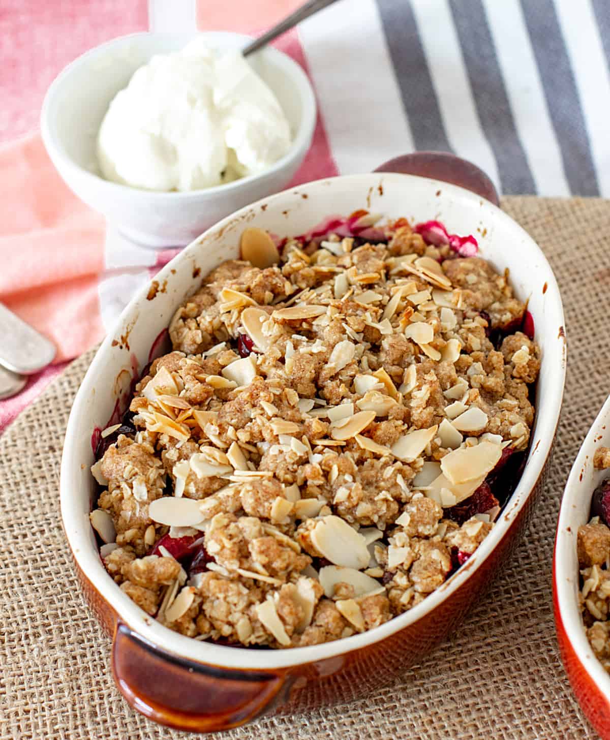 Striped red and blue cloth in the background with oval brown ceramic dish with cherry almond crumble. Burlap surface.