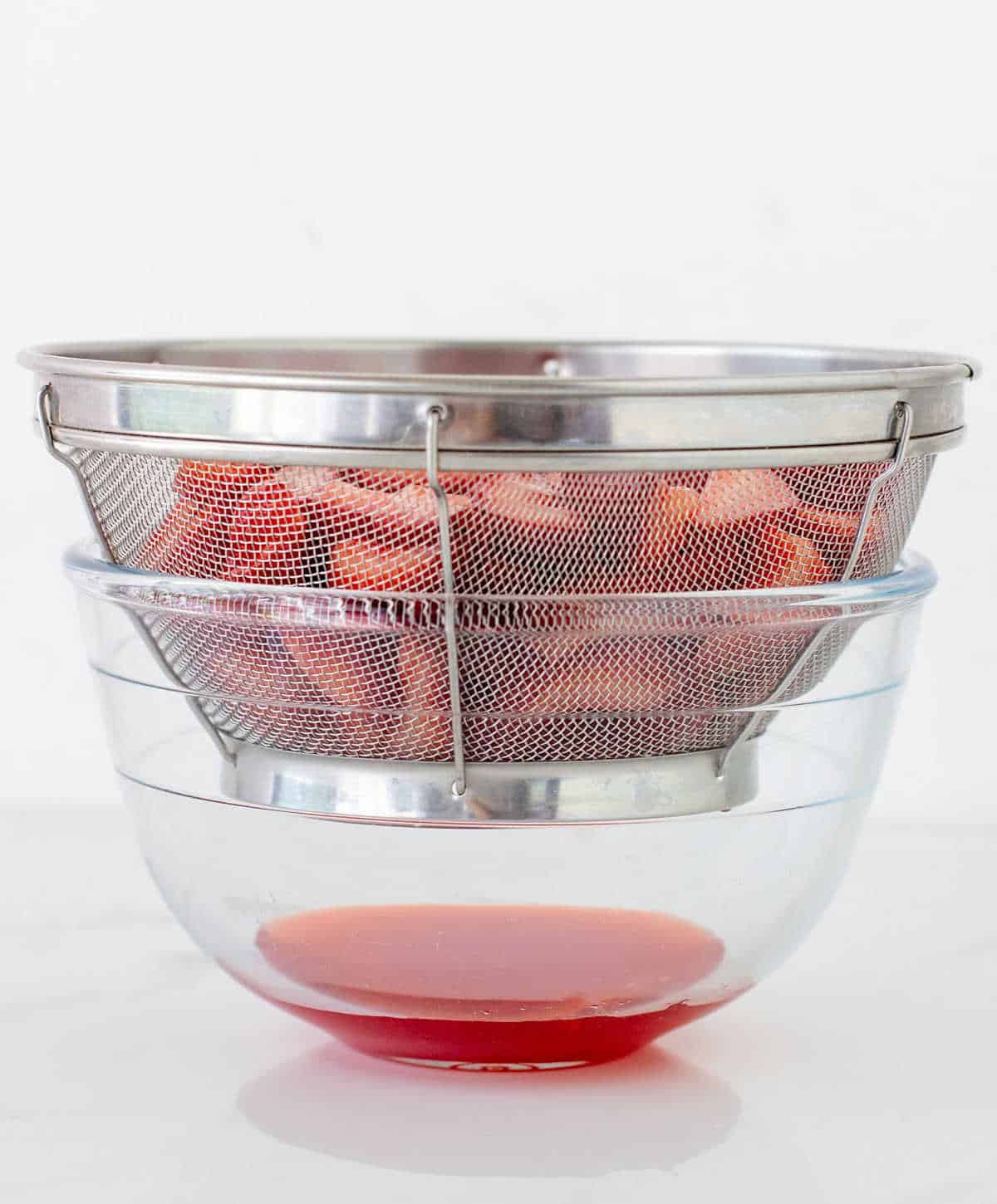 Colander with strawberries dripping juice over a glass bowl. White surface and background.