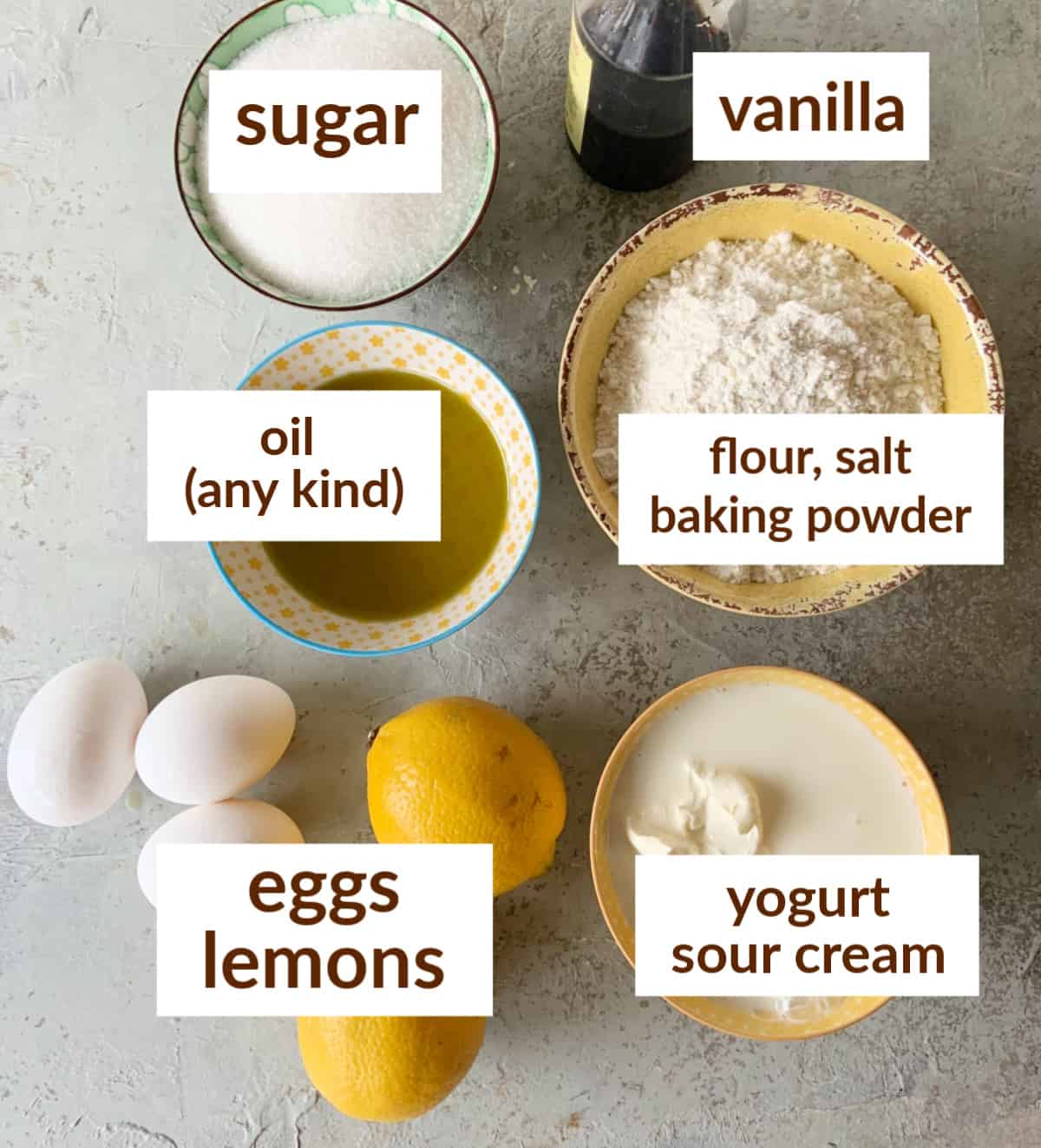Bowls on a grey surface with ingredients for lemon yogurt cake including eggs, oil, sugar, flour mixture, sour cream.