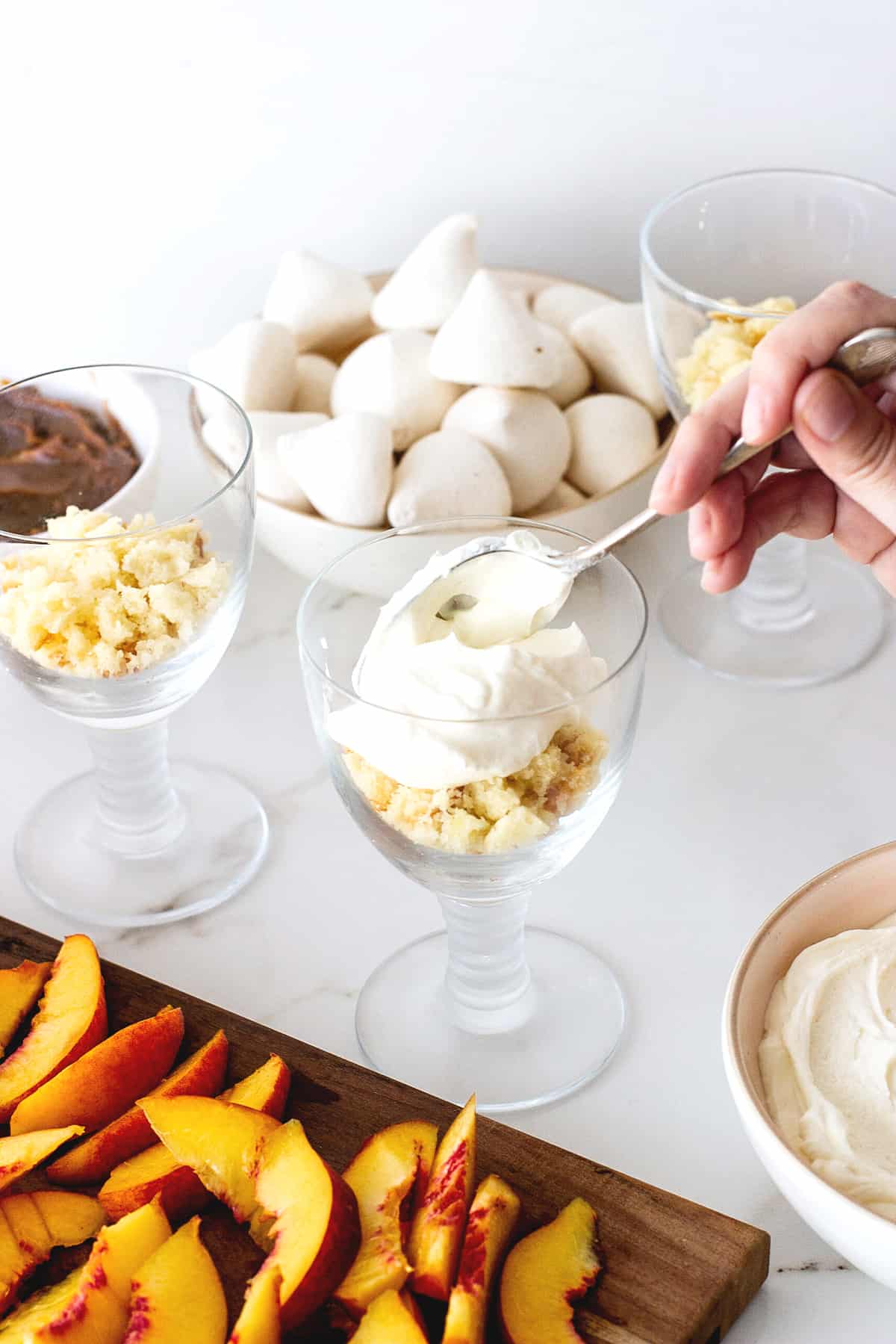 Hand adding cream to stem glass pn white surface; peaches, meringues, and rest of dessert around