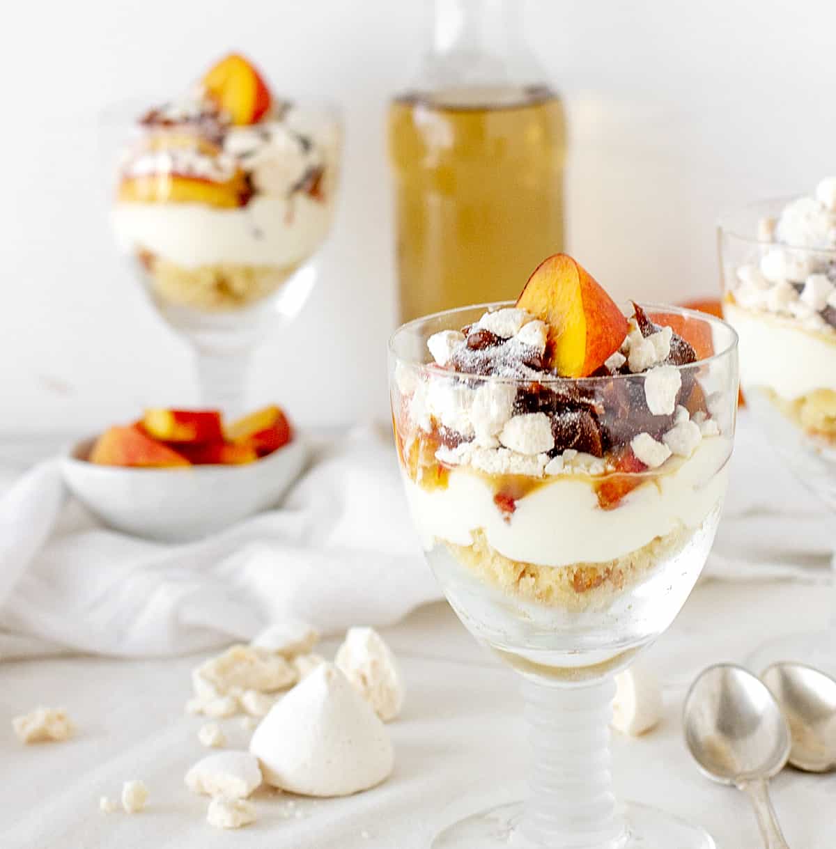 Long stem glasses with peaches and cream dessert, white surface, bottle and bowls in background.