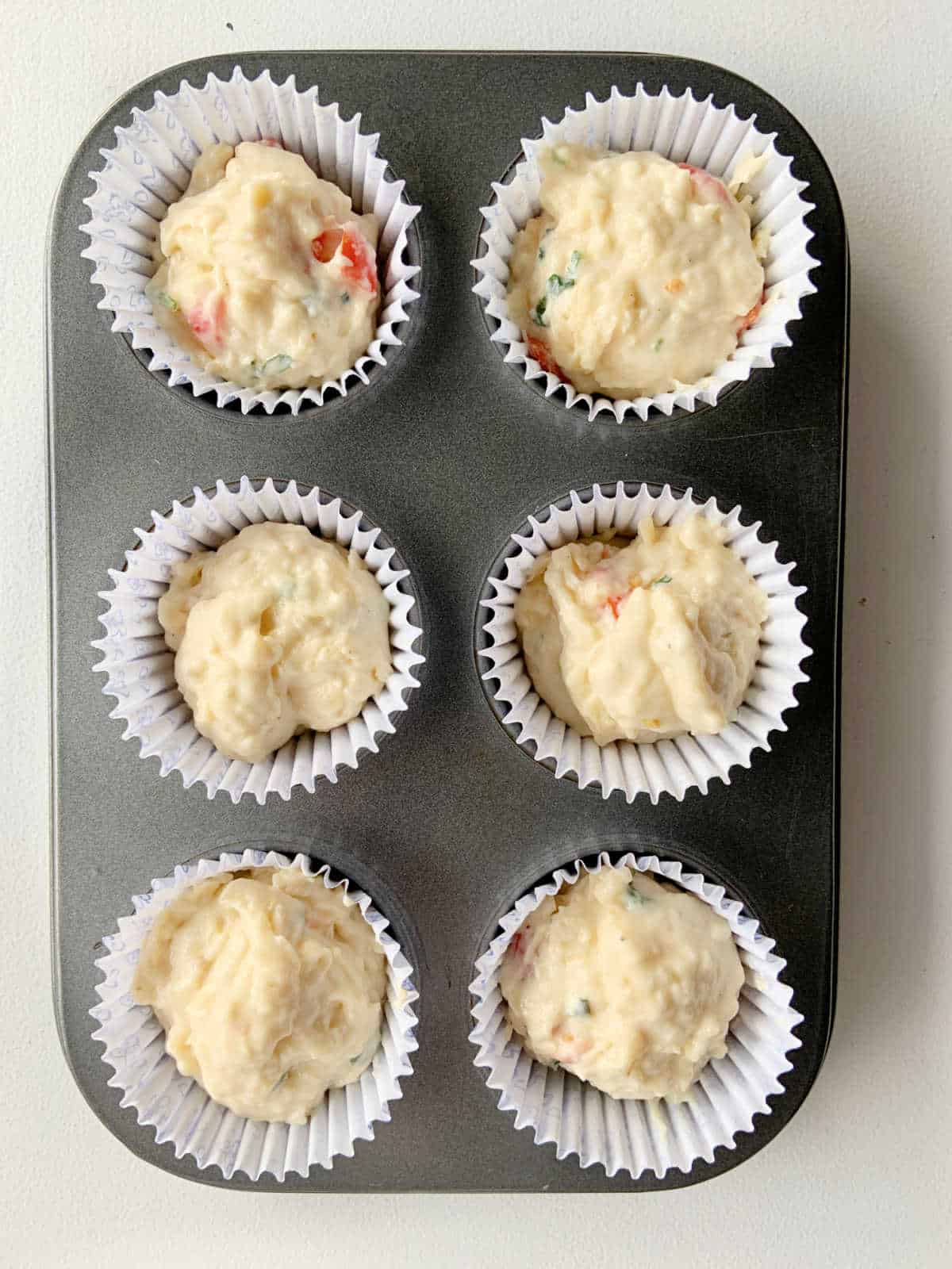 Muffin liners with savory cheese tomato batter in a metal pan on a white surface.