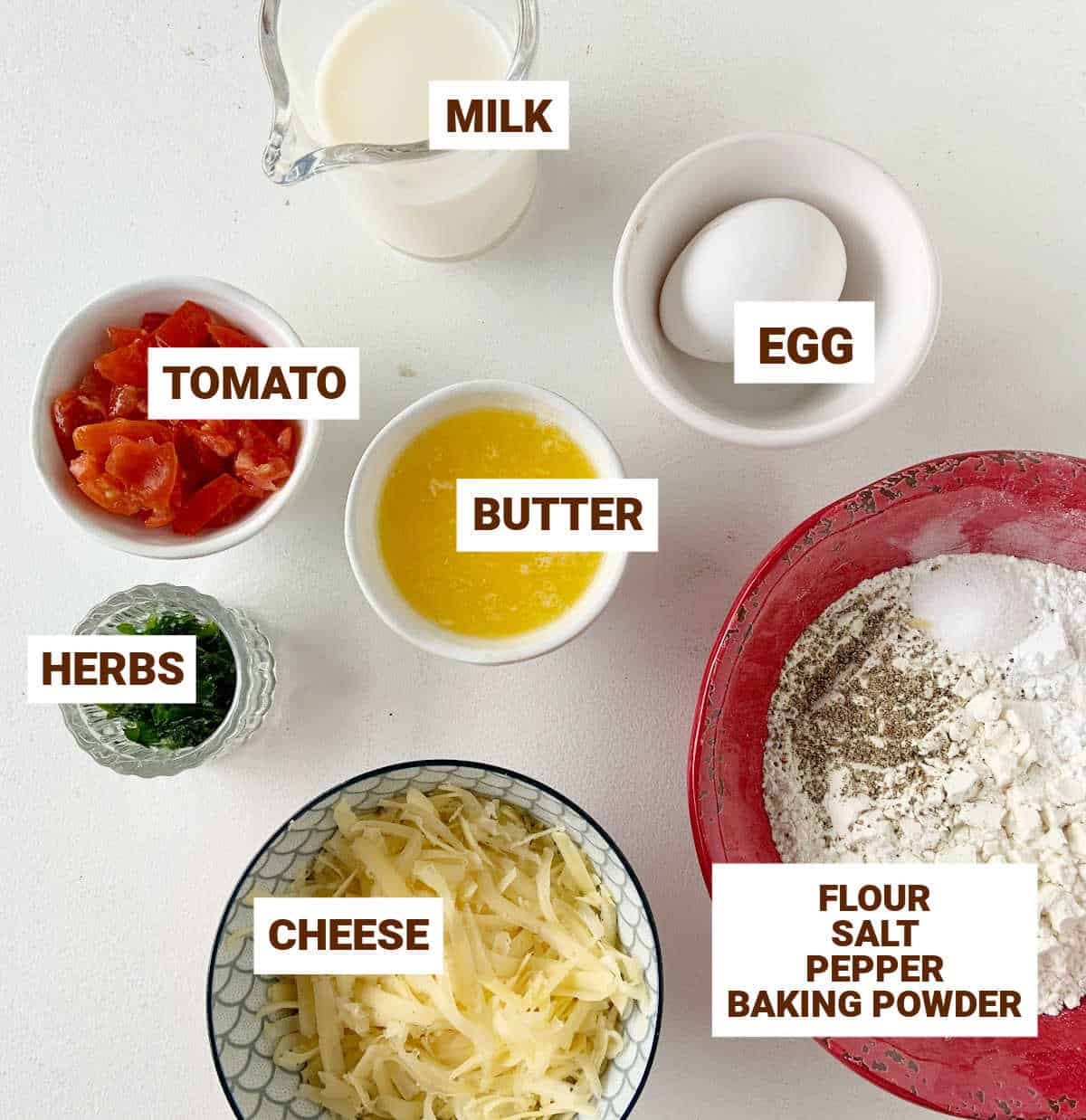 Bowls containing ingredients for savory muffins including cheese, tomato, sesonings, egg, flour mixture, milk, and butter. White surface.