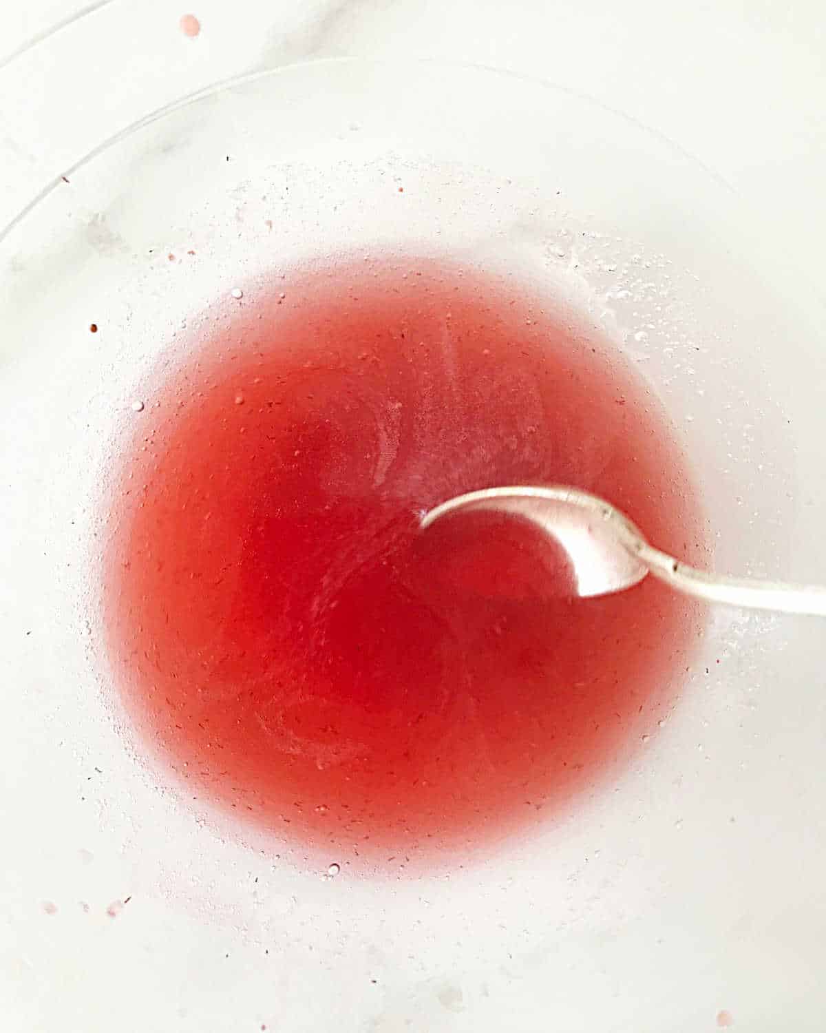 Stirring gelatin and strawberry juice with a silver spoon in a glass bowl on a white surface.