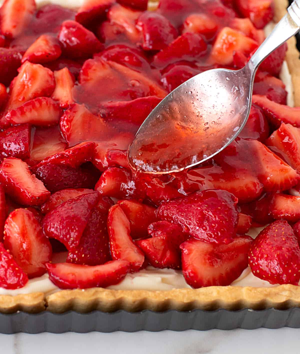 Adding strawberry jelly with spoon to tart topped with strawberries.