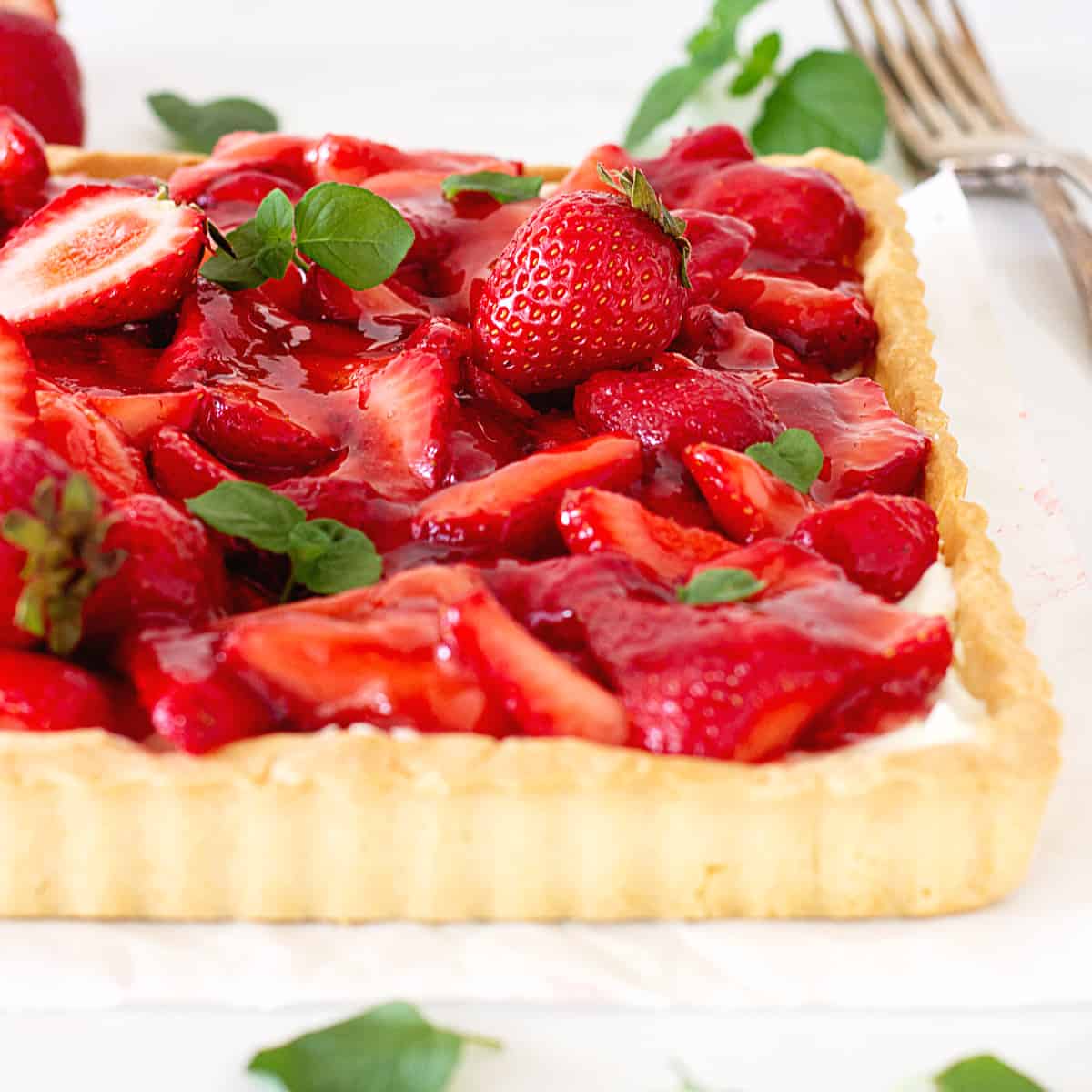 Partial slanted view of strawberry tart on a white surface, mint leaves on top.