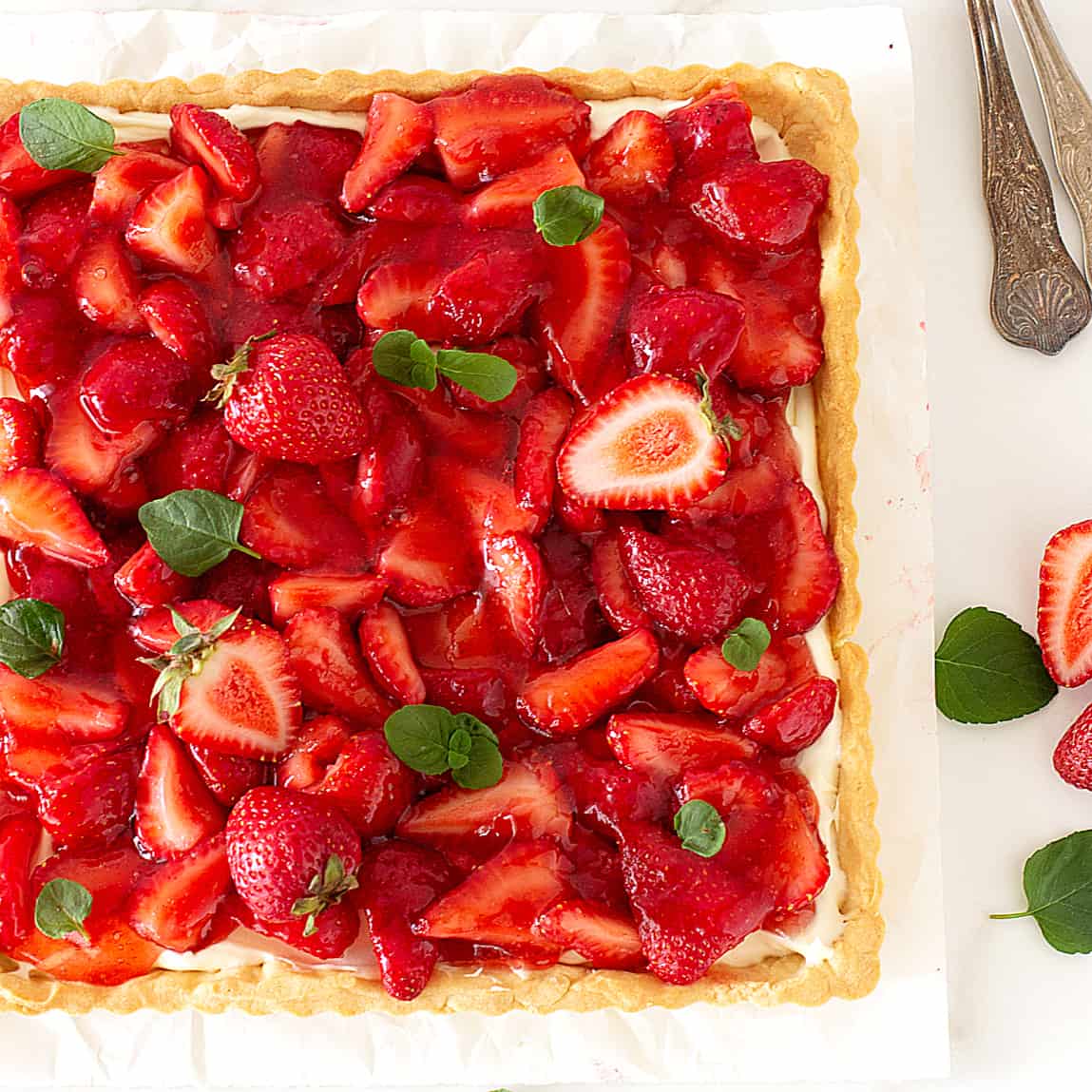 Partial view of strawberry tart on white surface, mint leaves and fork beside