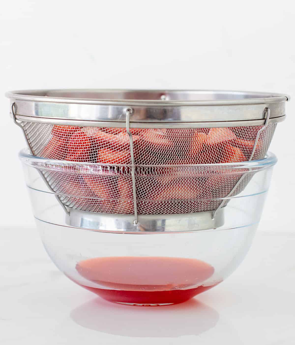 Metal colander with strawberries over a glass bowl, red juice dripping
