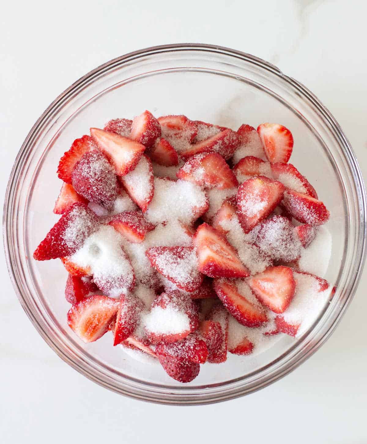 Glass bowl with fresh strawberry slices and sugar on a white surface.