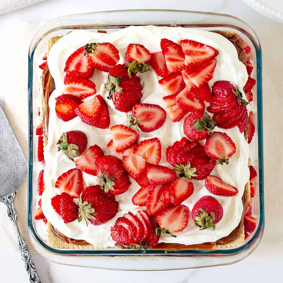 Glass square dish with strawberries and cream dessert; a silver cake server on the side.