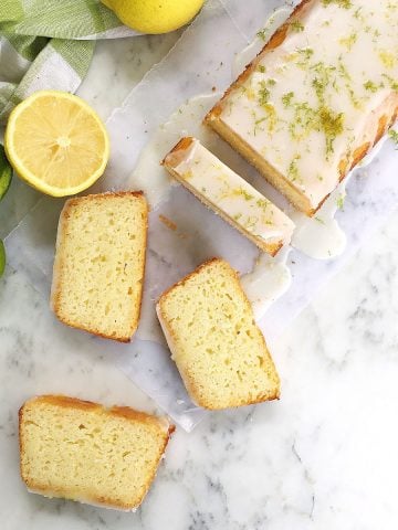 Top view of whole and sliced glazed citrus loaf cake, white marble surface, lemons and limes around.