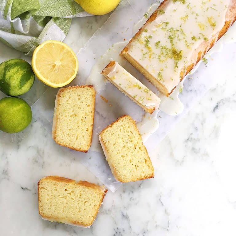 Top view of whole and sliced glazed citrus loaf cake, white marble surface, lemons and limes around.