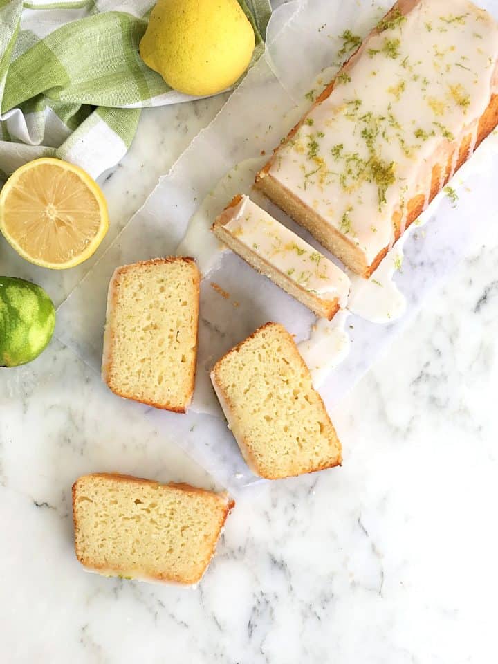 Overview of partial and slices of glazed loaf cake, white marble surface, lemons and limes nearby