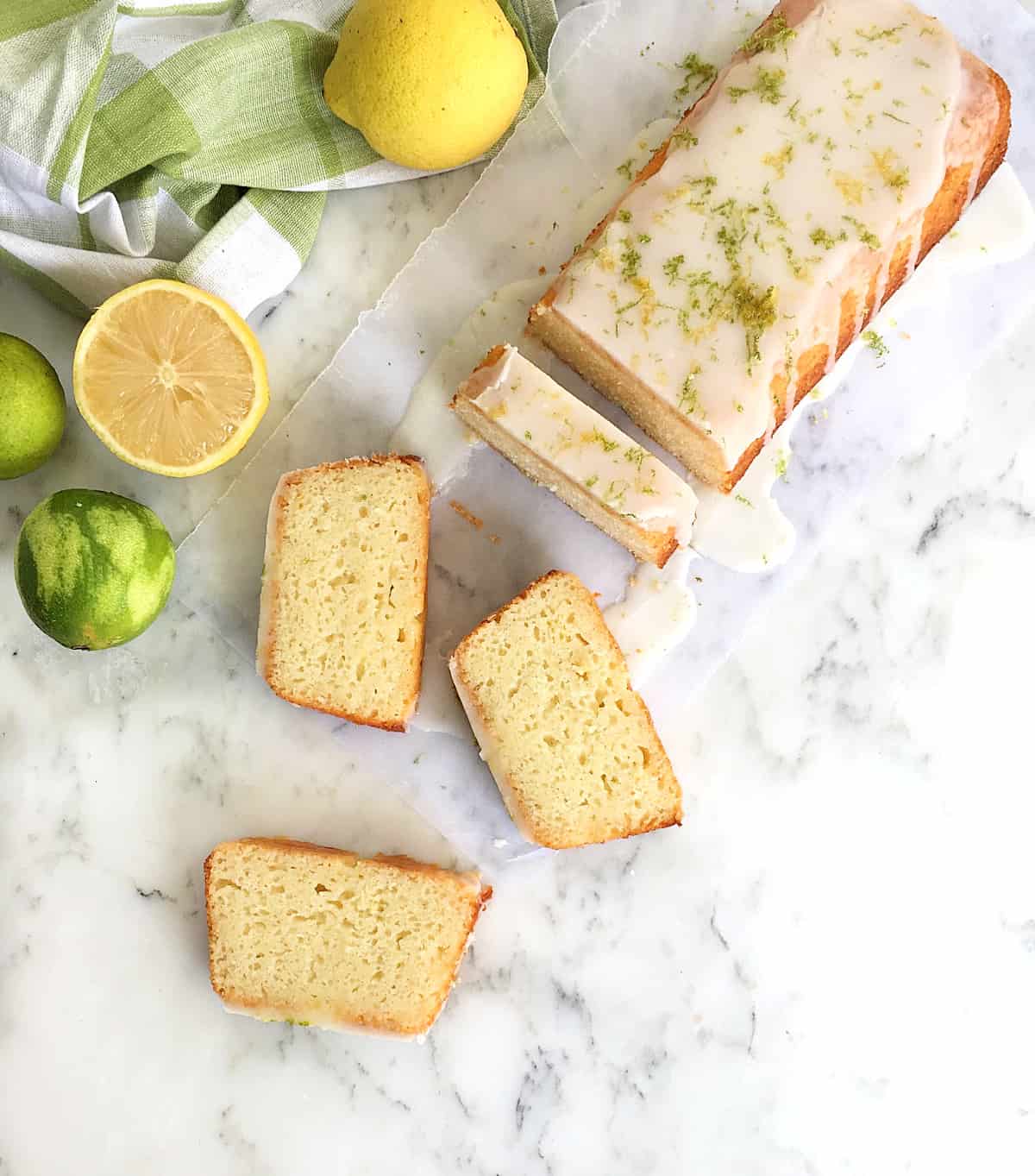 Overview of cut loaf cake, three slices, lemons and limes around, marble surface