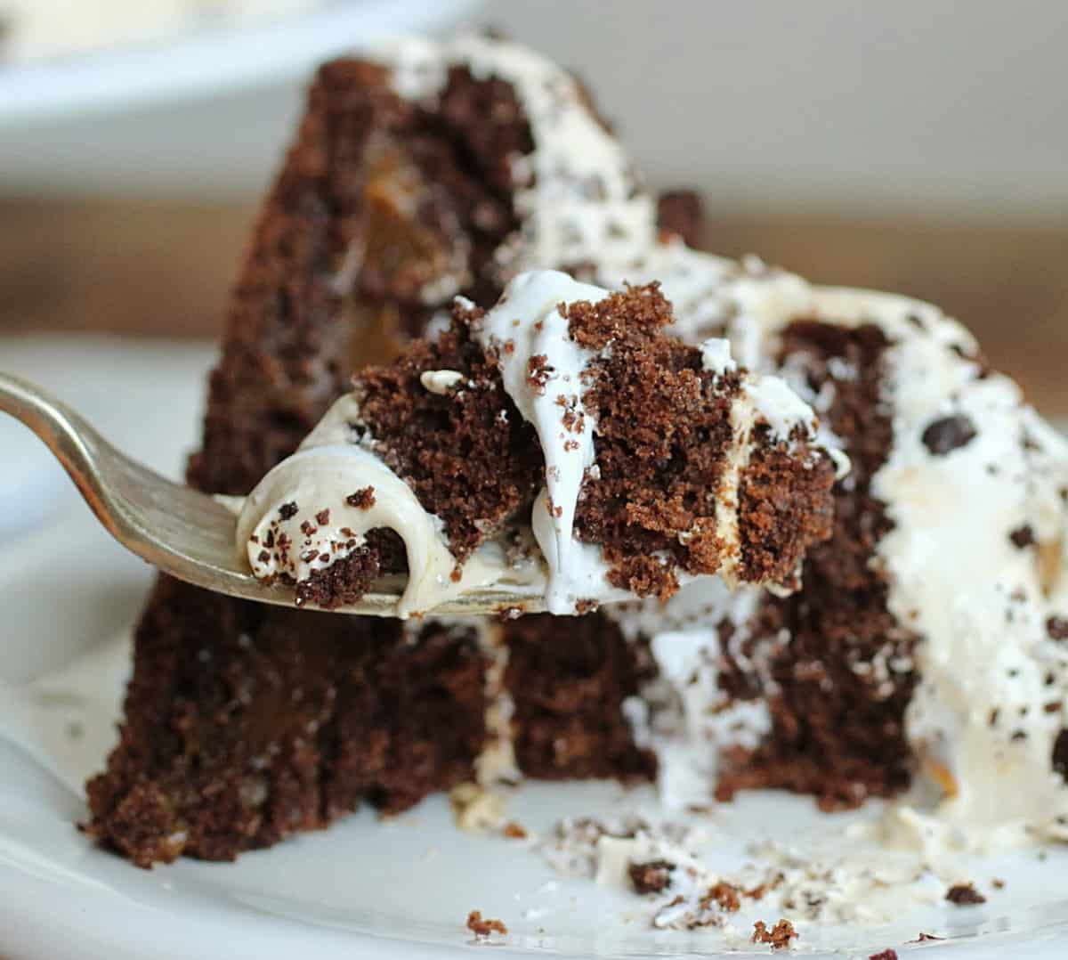 Silver fork with bite of chocolate layer cake with meringue, cake slice on white plate.