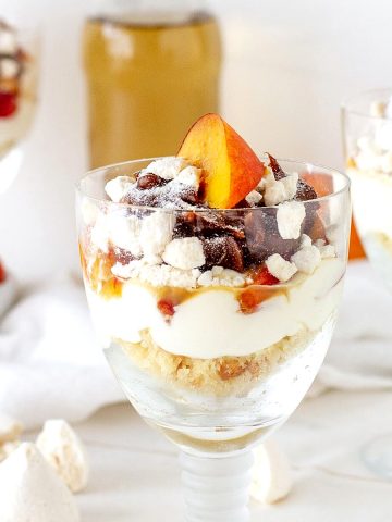 Several glasses with peach trifle servings, a white surface, bottle with white wine in the background.