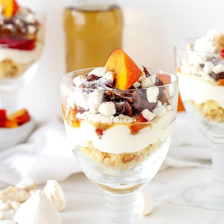 Several glasses with peach trifle servings, a white surface, bottle with white wine in the background.
