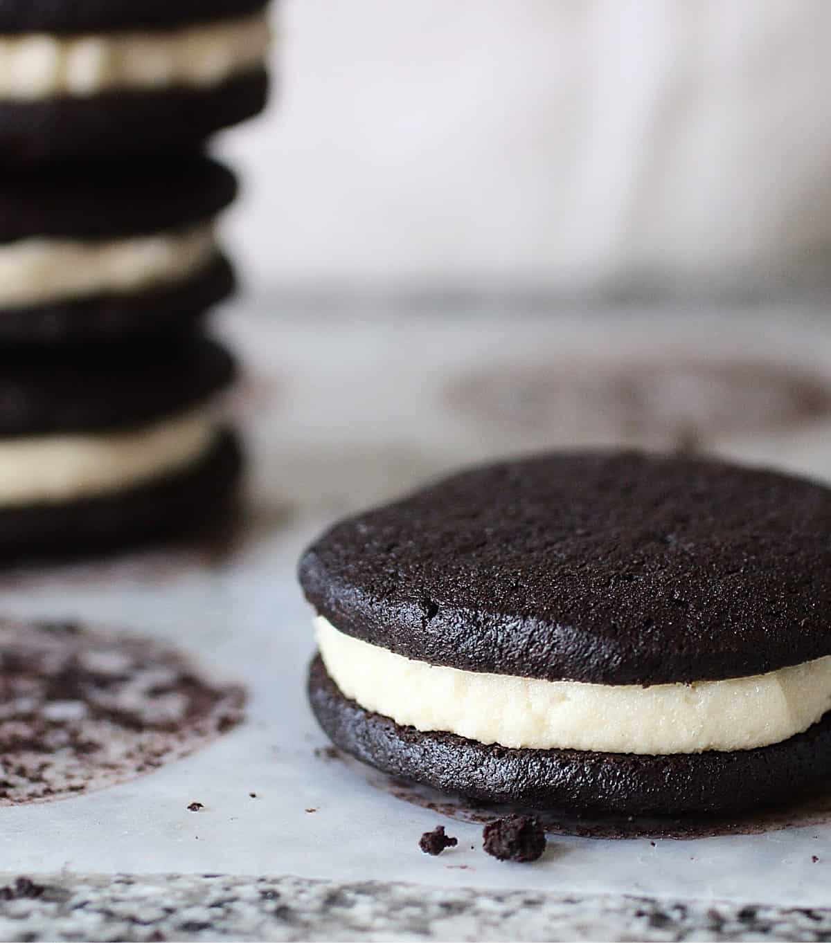 Single dark chocolate sandwich cookie on grey surface; stack of cookies in background